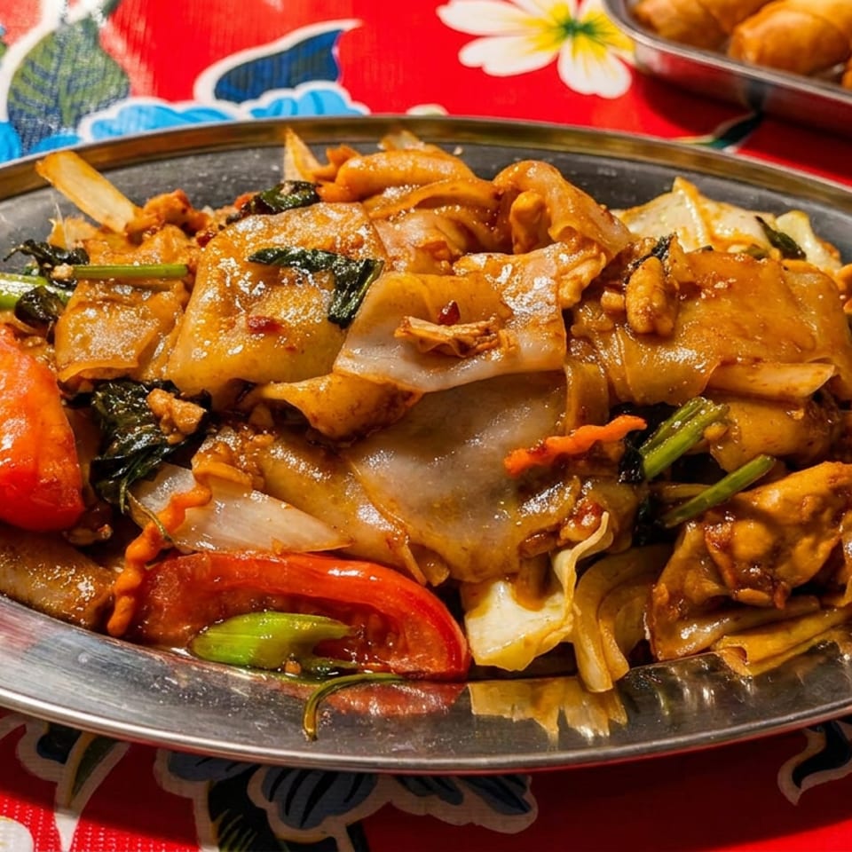 Plate of saucy stir-fried noodles with vegetables and chicken on a colorful floral tablecloth, with spring rolls in the background.